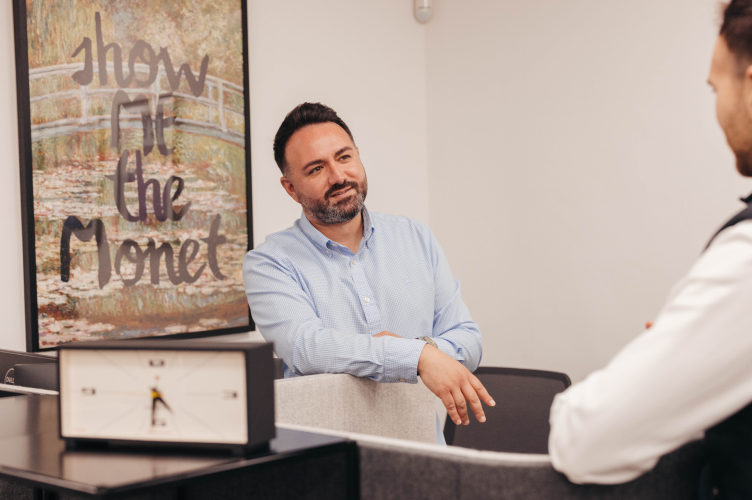 A man in a blue shirt engaged in conversation, standing in an office with a 'Show Me the Monet' art piece in the background.