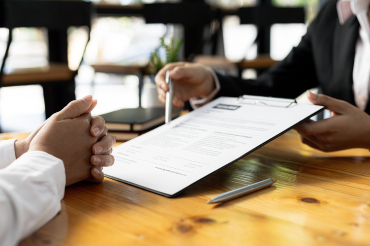 Two individuals reviewing a loan application document together at a wooden table, with one person holding a pen and pointing at details on a clipboard.