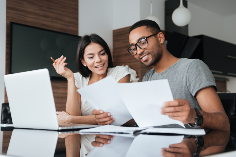 Two people sitting at a table, collaborating over documents while using a laptop. They appear to be smiling and engaged in discussion, suggesting a productive and positive working environment.